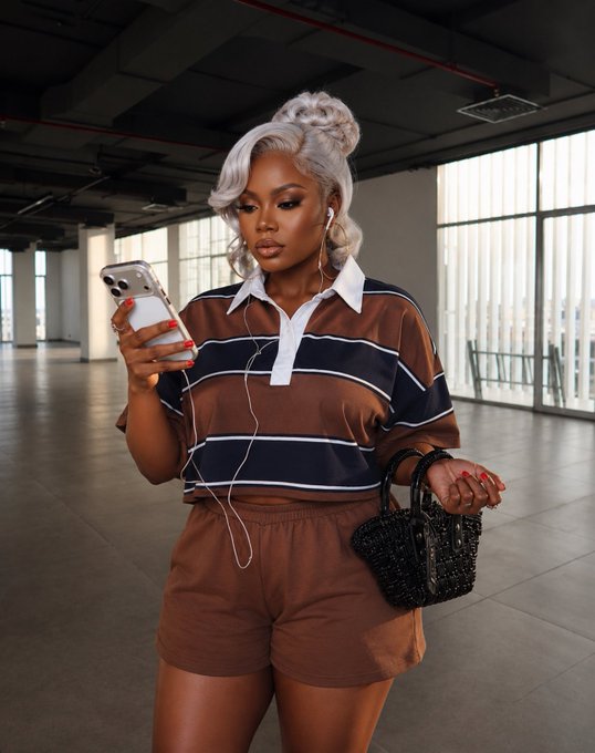 A stylish woman with platinum blonde hair and red nails looks down at her phone while wearing a brown and navy striped polo shirt with matching shorts, holding a black beaded handbag. The image reflects the post's casual, on-the-go vibe as she appears to be checking her phone—perhaps hoping to be noticed by @kosgoood—during a busy day before signing off for the night.