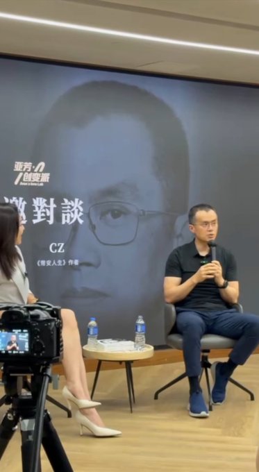 A man speaks into a microphone during an interview while a woman sits across from him, with a large backdrop displaying a portrait and the text "亞洲創變者" and "CZ" (likely referring to Changpeng Zhao of Binance). The image captures the moment described in the post, showing CZ's notable presence in the interview setting.