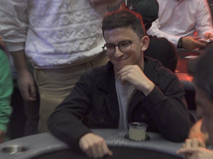 A young man with glasses and a black jacket smiling at a poker table, holding a hand of cards near his face in a dimly lit casino setting.