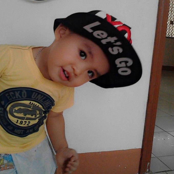 A young boy wearing a yellow Ecko Unltd t-shirt and a sideways black baseball cap that reads 'Let's Go', posing for the camera indoors.