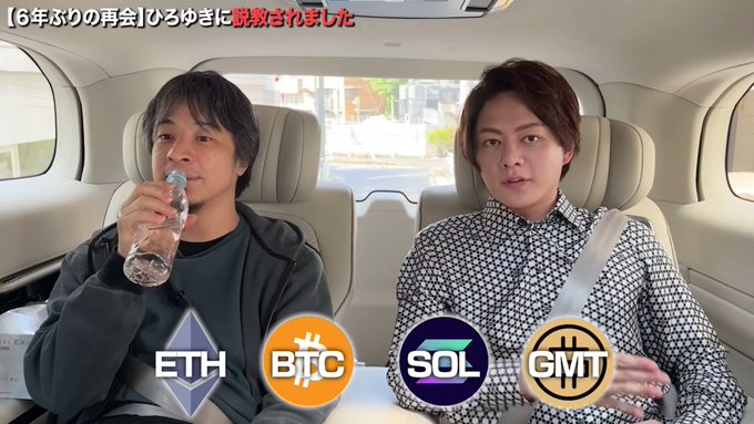 The image shows two men sitting inside a car, with one man drinking from a water bottle while the other man looks at him. The background of the image features logos for Bitcoin, Eth, and Sol, suggesting that they are discussing or comparing these digital currencies during their conversation in the car.