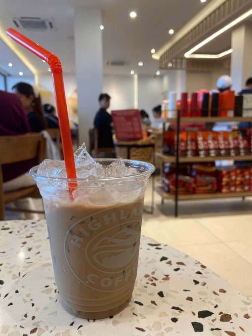 A cup of Highlands Coffee iced coffee with a red straw sits on a terrazzo table in a café, with shelves of coffee products and other customers visible in the background—matching the post's message about finally recharging with coffee for a productive work day.