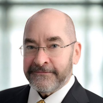 A professional headshot of a bald man with a gray beard wearing glasses, a dark suit, and a yellow tie.
