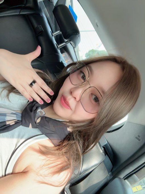 A young woman with glasses and long hair takes a selfie in a car, showing her hand with pink nails and a ring.