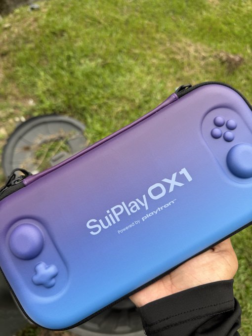 A hand holds a gradient purple-to-blue "SuiPlay OX1 powered by Playtron" gaming handheld in front of a grassy outdoor setting, fitting for a post about Sui Basecamp Singapore ending—likely capturing a moment of relief or celebration with a new gadget in hand.