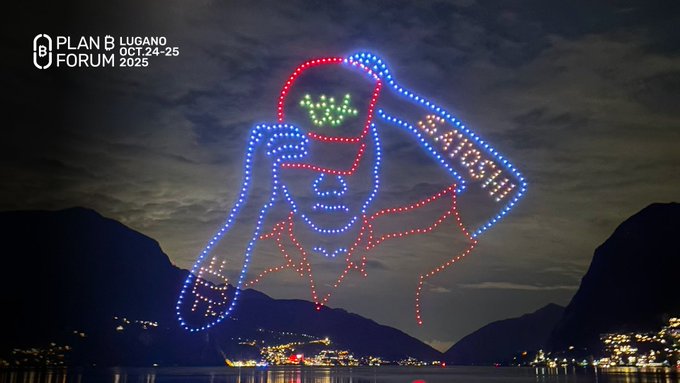 A large illuminated drone show depicts a person with green glasses and blue/red lights, displayed over a lake at night for 'PLAN B LUGANO FORUM OCT 24-25 2025'.