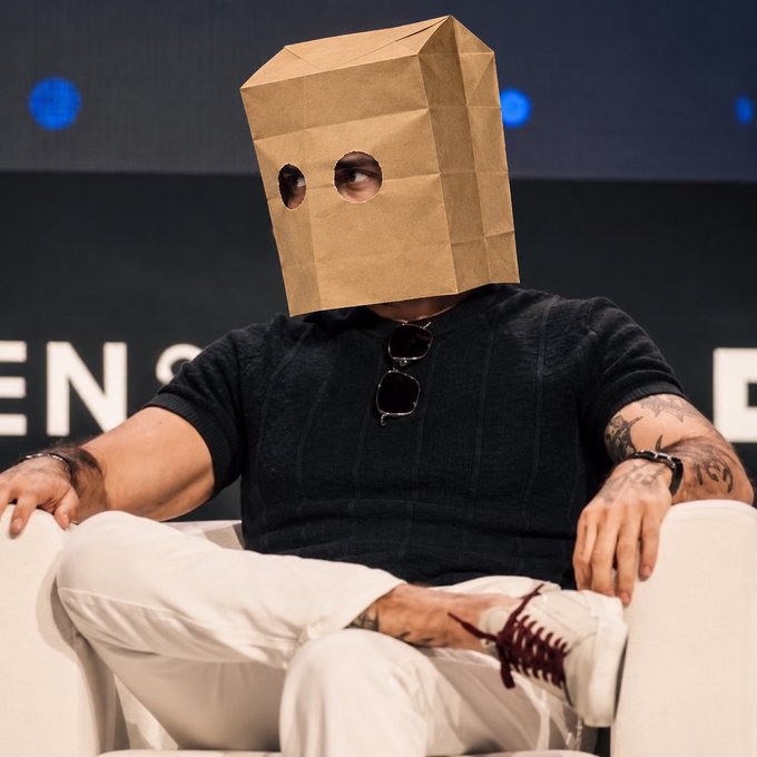 A photograph of a man sitting in a white chair wearing a brown paper bag over his head with holes cut out for his eyes, dressed in a black sweater and white pants with visible tattoos on his arms.