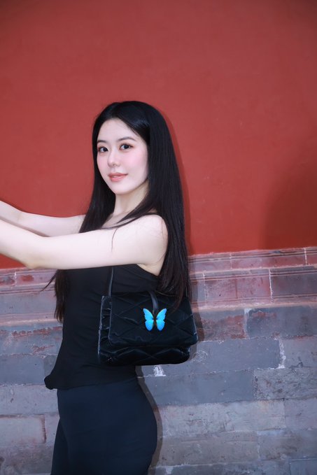 A confident woman with long dark hair poses against a red wall and gray brick base, holding up a black quilted handbag adorned with a striking turquoise butterfly emblem that visually echoes the "Flap" brand identity mentioned in the post.