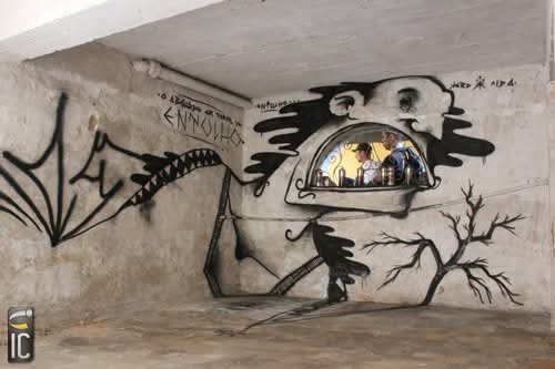 A graffiti mural of a surreal creature with an open mouth containing a small scene, along with other black spray-painted designs, on a concrete wall.