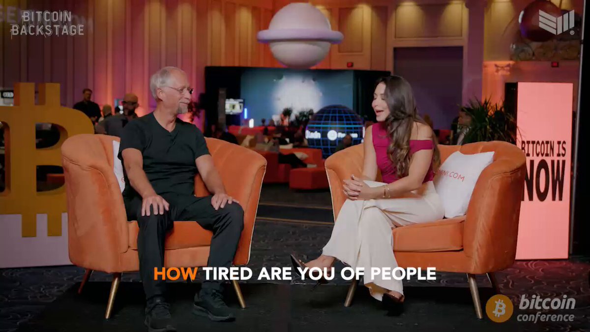 The image shows two men sitting in chairs at a conference room with a large screen behind them, which displays an interview between the hosts of the Bitcoin conference. The hosts are discussing the topic "How tired are you of people?". One man is seated on an orange chair while the other is on a white one. They are talking about how tired they are and how it affects their daily lives.