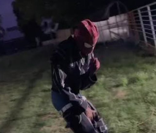 A nighttime photo of someone wearing a Spider-Man mask and a black costume, posing in a grassy backyard.