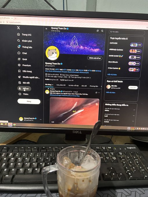 A Dell computer monitor displays the X (formerly Twitter) account 'Quang Tuam Do' with EOS branding, while a glass mug with iced coffee and a spoon sits on the keyboard in the foreground.