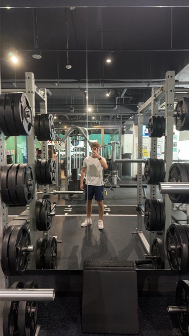A person takes a mirror selfie with their phone in a gym weight room, surrounded by barbells and weight plates.