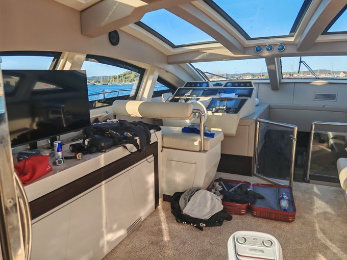 The interior cabin of a luxury yacht is shown, featuring a helm station with controls, a mounted TV, scattered luggage, and a view of the water through the windows.