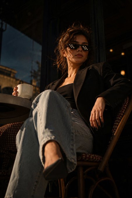 A woman wearing pixelated checkered sunglasses sits on a chair holding a cup.