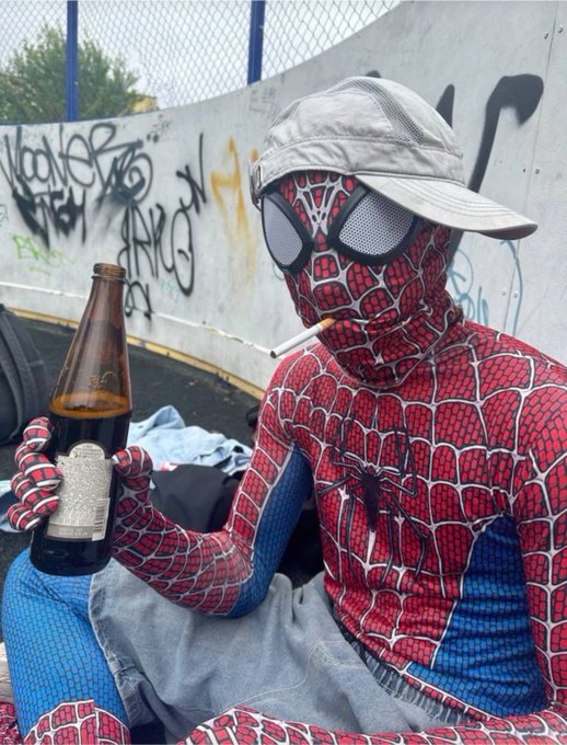 A person dressed in a Spider-Man costume and silver cap, wearing large sunglasses, sitting outdoors against a graffiti-covered wall while holding a beer bottle and a cigarette.