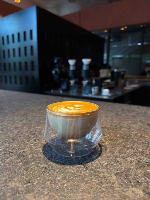 A photograph of a cappuccino with latte art served in a double-walled glass cup on a speckled stone countertop, with espresso equipment visible in the background.