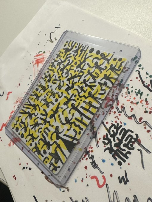 A mixed-media artwork features a yellow cassette tape covered in dense black scribbled markings, resting on a white surface splattered with colorful paint drips and graffiti-style tags including what appears to read "GYM."

This image relates to the post as it showcases the artist's branded "Alien Inks" creative work, serving as an example of the original art they are promoting while seeking advice on the business side of being an artist.