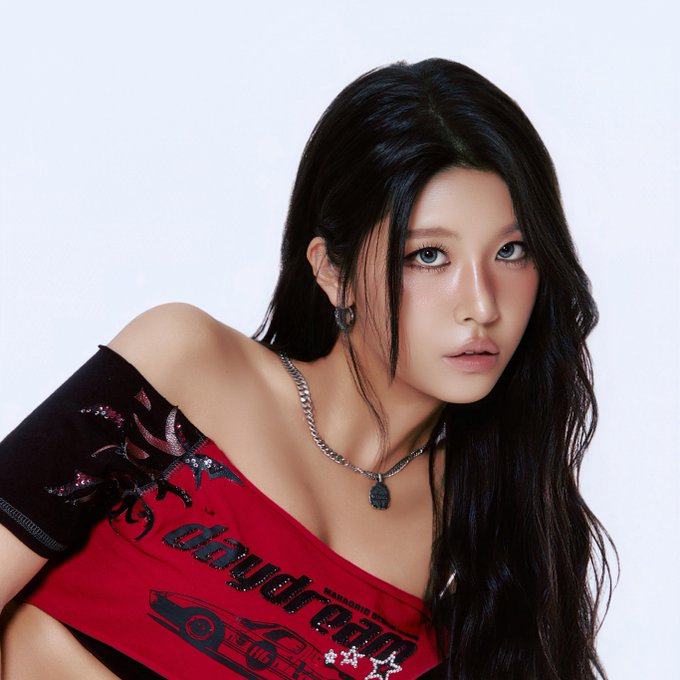 A young woman with long black hair poses wearing a red one-shoulder top with graphic prints and a black mesh sleeve.
