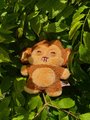 A small plush toy resembling a cartoon lion or cat character rests among green leaves.