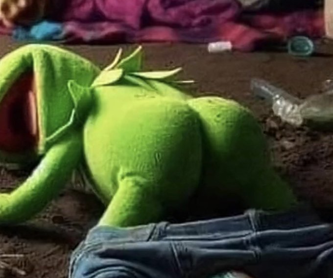 A green plush Kermit the Frog toy lies face-down on the ground next to a pair of blue jeans, with various debris and bottles scattered around the dirt area.