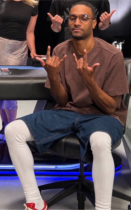 A man wearing glasses, a brown t-shirt, denim shorts over white leggings, and red sneakers, sitting in a chair and making hand gestures.