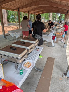 The image shows a group of people gathered under a pavilion, enjoying food and drinks together. There are several tables set up with trays of hot dogs and other dishes, indicating that they are having a picnic or a casual gathering. The attendees include both men and women, all dressed in casual attire. Some individuals can be seen standing around the tables, while others are seated at the tables enjoying their meal.

The post features a photo of a man wearing an @BoredApeYC shirt, which suggests that this event is related to a meetup or gathering for people with similar interests. The image also includes text about a Staple x BAYC shirt and a hot dog, further indicating the theme of the event. Overall, it appears to be a fun and relaxed outdoor social gathering where everyone can enjoy good food and company.