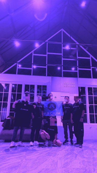 The image features a group of six people posing for a photo, all wearing black t-shirts with the word "ARCELERATE" printed on them. They are standing outside a building that has large windows and a glass roof, giving an impression of a modern architectural design. The group is positioned in front of a couch, which adds to the casual atmosphere of their gathering.