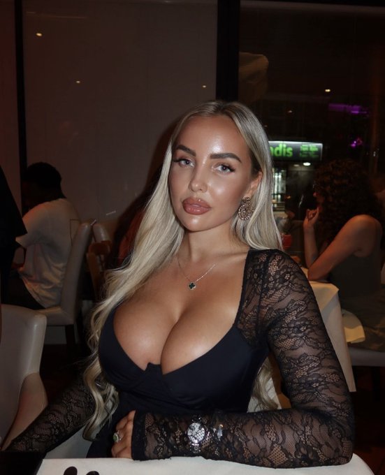 A blonde woman wearing a black lace-sleeve top poses for a portrait in a restaurant setting.