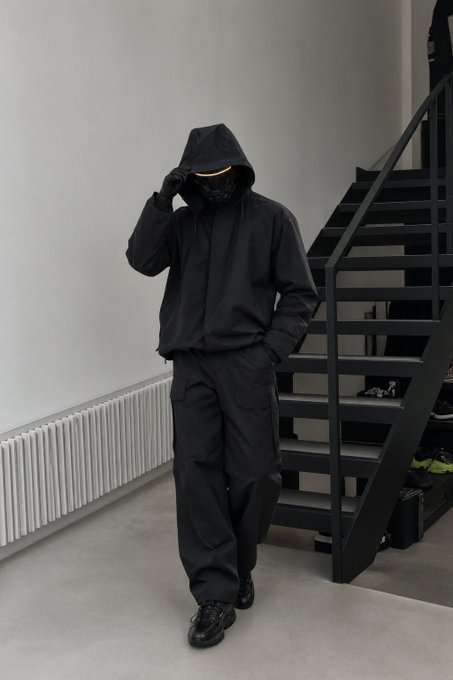 A person modeling a full black techwear outfit, including a hooded jacket, mask with a glowing visor, cargo pants, and chunky sneakers, standing on a staircase.