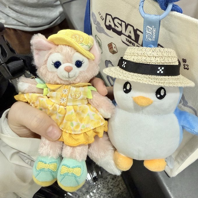 A plush LinaBell (pink fox in a yellow citrus-themed dress) and a plush penguin wearing a straw hat are held up in front of a tote bag reading "ASIA," with the penguin's tag showing "Peter Pan" — capturing the "Pengu" crypto boss and his "Xiao Mei" companion as they prepare for their trip to Shanghai.