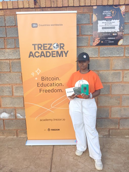 A young woman wearing an orange shirt and white pants holding a Trezor device, standing next to a 'Trezor Academy' banner advertising Bitcoin education across 28+ countries.