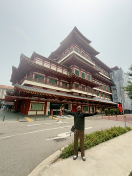 A person stands on a sidewalk posing in front of the multi-tiered Buddha Tooth Relic Temple in Singapore, a striking Chinese Buddhist architectural landmark with ornate red and gold details. The grand temple against the cityscape captures the cultural beauty referenced in the "Beautiful Singapore" caption.