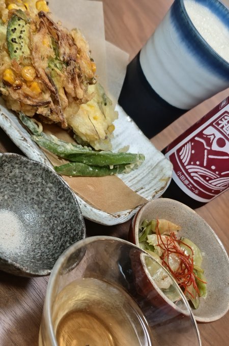 A Japanese meal featuring tempura vegetables on a plate, a small dish with what appears to be avocado and red threads, a bowl of rice, and a glass of an amber beverage.
