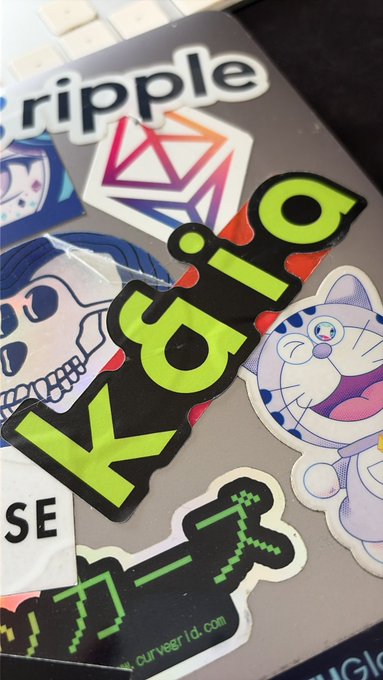 The image shows a laptop with various stickers, including one that reads "KAIA", placed on top of it. The stickers are colorful and diverse, adding character to the laptop's exterior. The text "$KAIA" stands out prominently in the image, suggesting that this particular sticker might be related to an event or promotion involving the name "Kaia".