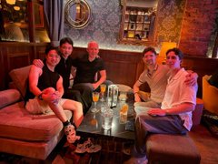 A group of five people gather around a table with drinks in a dimly lit lounge, posing and smiling for a photo as they spend time together late at night. The image captures the casual, friendly moment referenced in the post about getting outfit advice from the Monad team at 11pm.