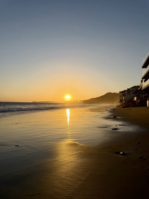 A serene sunset over a beach with golden light reflecting on the wet sand, houses visible along the shoreline on the right.