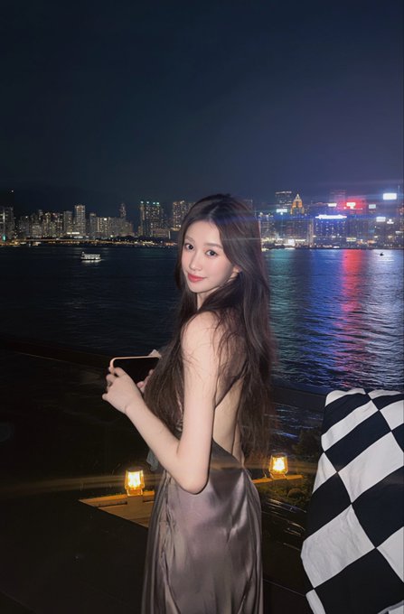 A woman in a gray satin dress holds a phone at a waterfront location at night, with a brightly lit city skyline across the harbor.