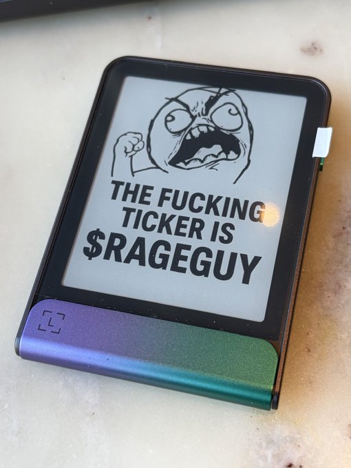 An e-ink display device with an iridescent green/purple casing showing a rage face meme with the text "THE FUCKING TICKER IS $RAGEGUY".
