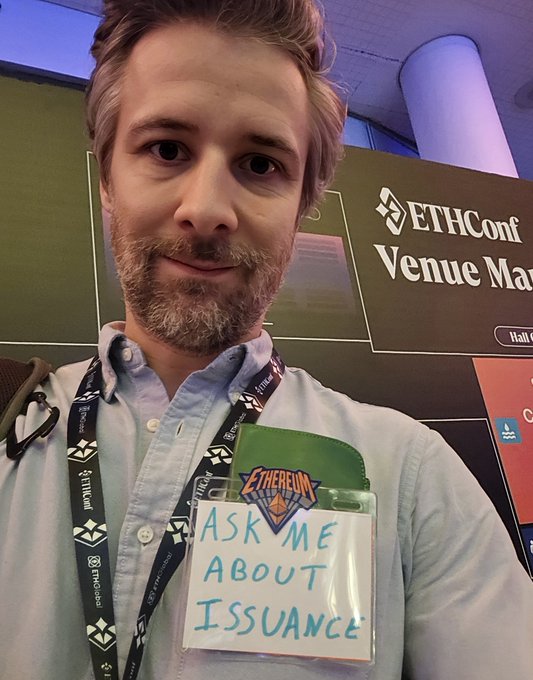 The image features a man wearing a lanyard with a name tag that reads "ask me about how Ethereum is going to print less and burn more". The man appears to be at an event, possibly a conference or exhibition, as indicated by the presence of a sign in front of him. The sign displays the words "ETH CONF" and "VENUE MARS", which suggests that this could be an Ethcoin event or convention.