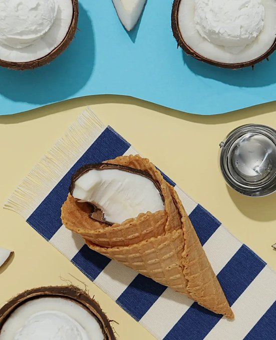A waffle cone filled with coconut cream sits on a blue and white striped napkin, surrounded by coconut halves and a silver ice cream scoop on a yellow and blue background.
