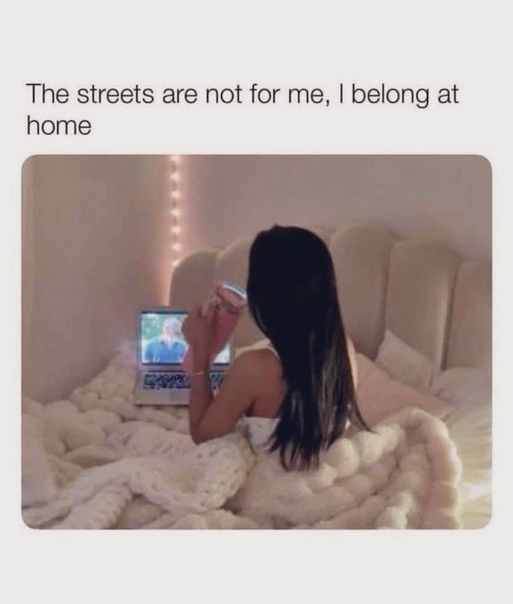 The image shows a woman lying down on her bed, using a laptop computer while holding a cup of coffee. The text above the photo reads "The streets are not for me I belong at home", which seems to be a humorous or satirical statement about the current situation.