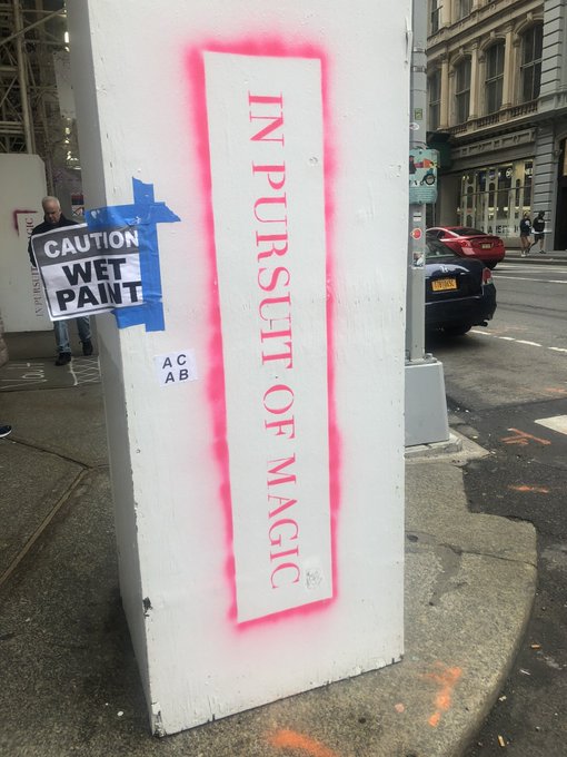An urban street scene features a white utility box with pink graffiti reading 'IN PURSUIT OF MAGIC', partially covered by a 'CAUTION WET PAINT' sign.