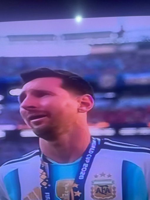 A close-up shot of Lionel Messi wearing an Argentina national team jersey and a FIFA World Cup medal lanyard, appearing emotional in a stadium setting.