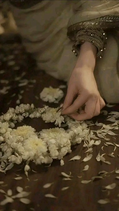 Artistic photo of a person wearing an ornate beaded bracelet and flowing fabric, delicately arranging white chrysanthemum flowers on a wooden surface scattered with petals.