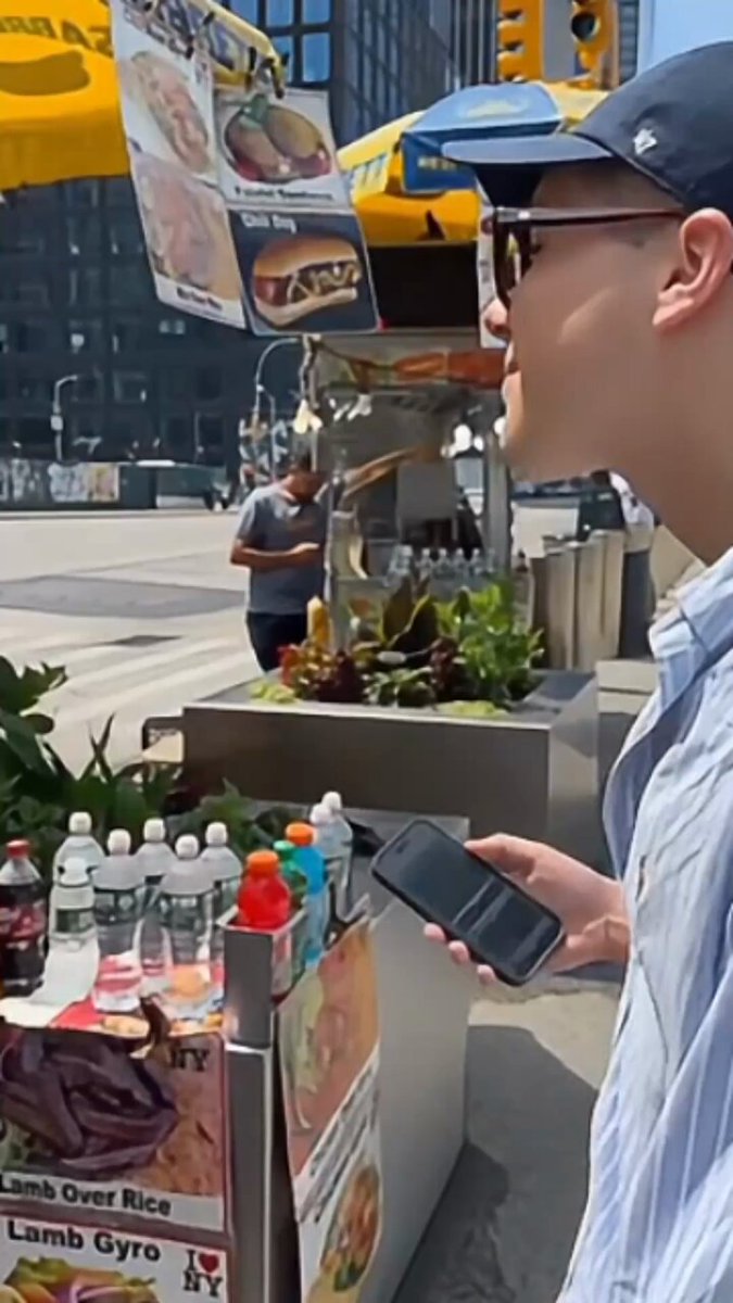 The image shows a man standing next to a food cart selling hot dogs while using his cell phone. The food cart has an advertisement for lamb gyro, which suggests that the location might be near a restaurant or food establishment serving this dish. The man is holding a wallet and appears to be paying with it, possibly using a mobile payment app like @MiniPay Card.