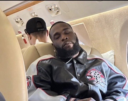 A bearded man in a black leather jacket with red trim relaxes with his eyes closed in a private jet cabin, while another passenger sits behind him.