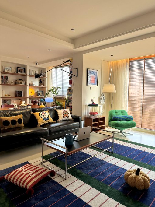 A cozy, well-decorated living room featuring a black leather couch with character pillows, a glass coffee table with a laptop, a green textured chair, a colorful striped rug, and built-in shelves filled with figurines and books.