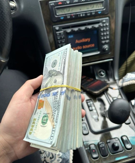 A hand holds a thick stack of $100 bills banded with a rubber band inside a car, with the dashboard display reading "Auxiliary Audio source," illustrating the "making money" part of the weekend hustle before stepping outside.