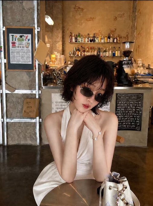 A young woman with short dark hair and small oval sunglasses poses thoughtfully at a table in a cozy café, wearing a white halter dress and a delicate bracelet. Behind her, a chalkboard menu lists coffee options like "FILTER COFFEE 60B" and "HOT CHOCOLATE 80B," while shelves of liquor bottles line the wall—creating a stylish, curated backdrop that contrasts with the post's message, which promotes earning money through social media engagement schemes rather than legitimate work.